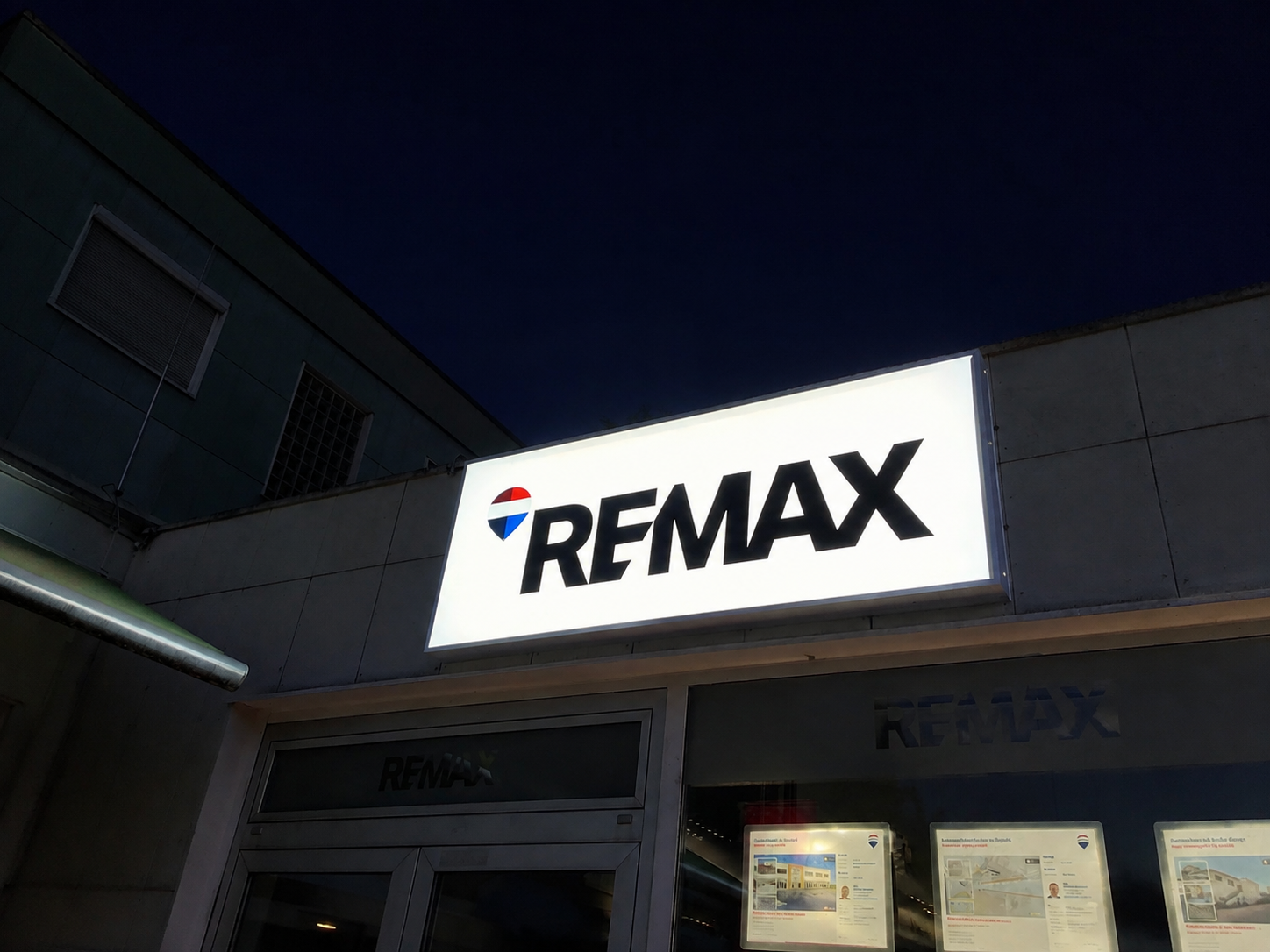 Großflächiger RE/MAX Leuchtkasten mit gleichmäßig ausgeleuchteter Acrylfront an einer Fassade bei Nacht