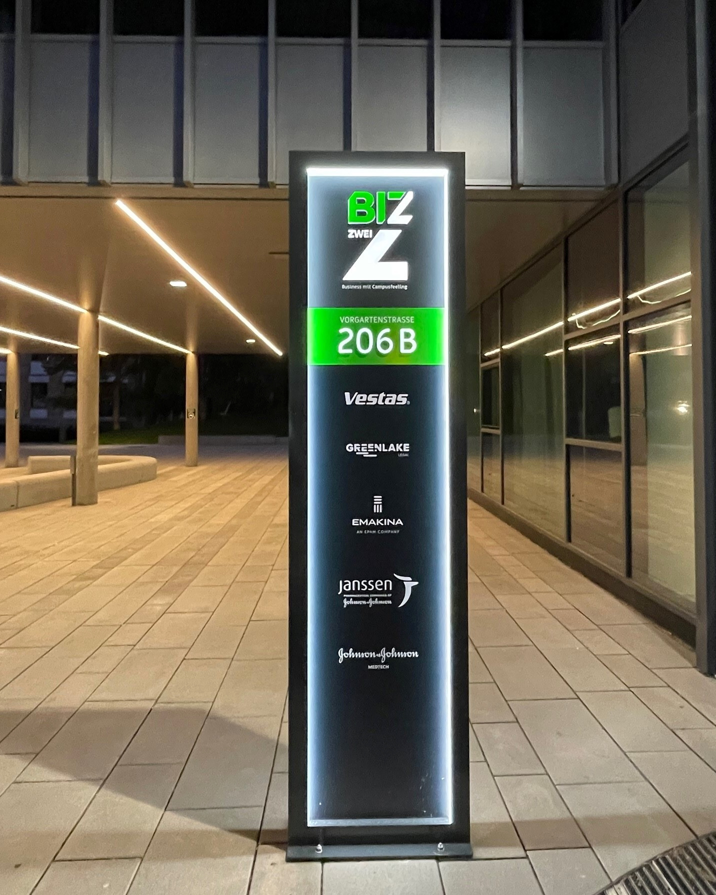 Beleuchteter BIZ-Pylon mit Gebäudeadresse und Unternehmenslogos vor einem Bürogebäude