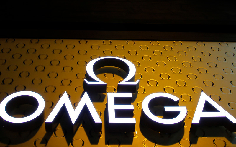 Beleuchteter OMEGA Schriftzug als superflacher Frontleuchter vor einer goldenen, mit Omega-Symbolen gemusterten Wand