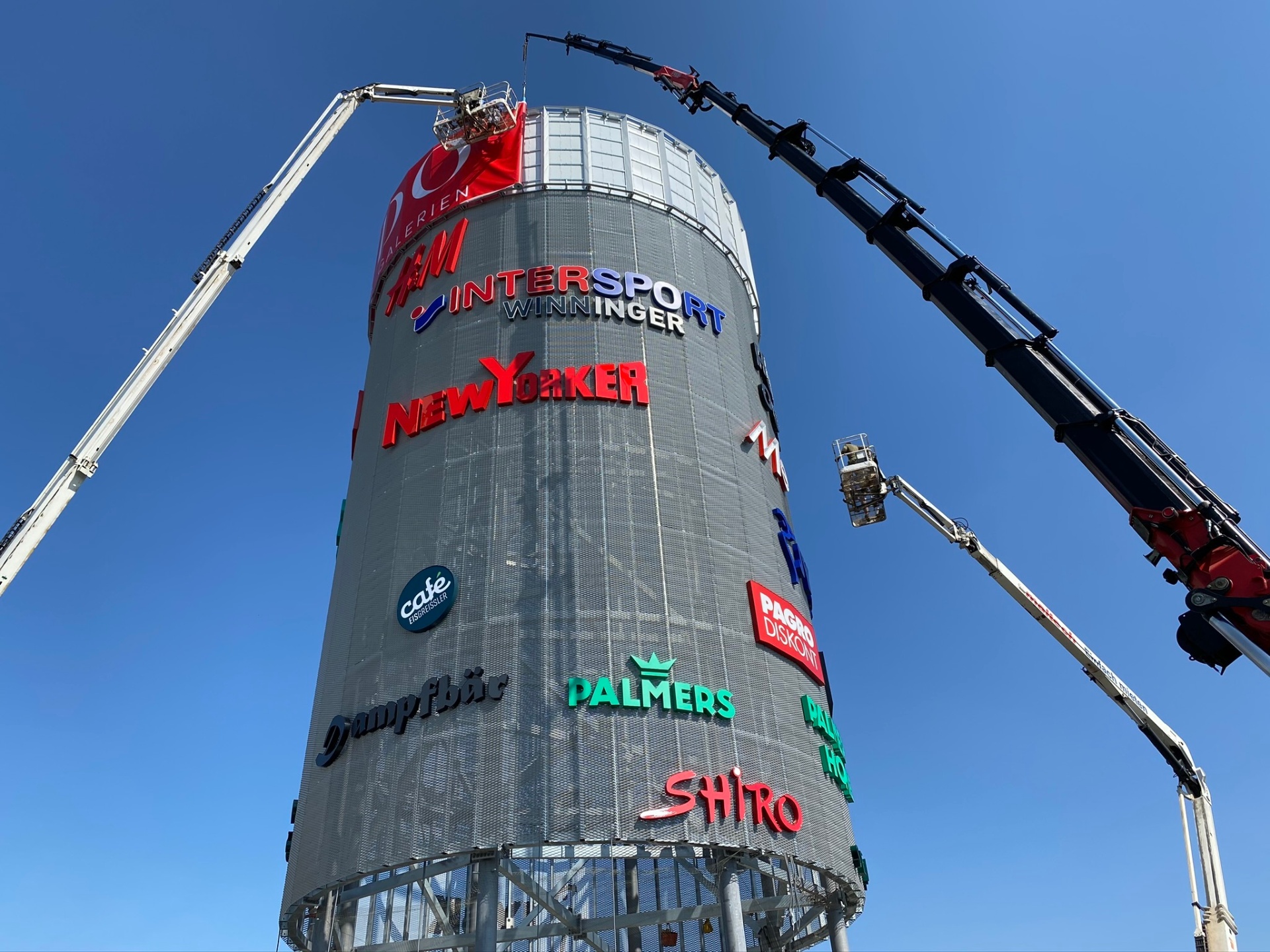 Zwei Hubsteiger im Einsatz an einem hohen Pylon mit zahlreichen Markenlogos