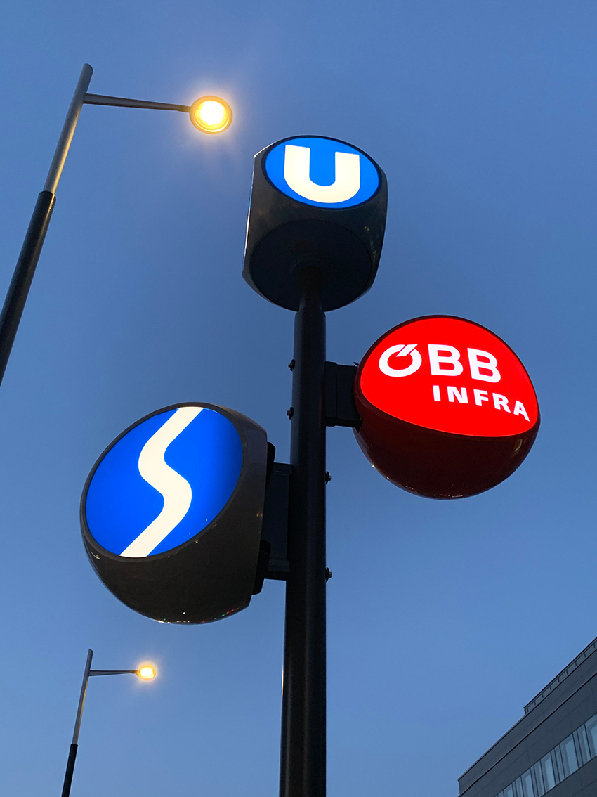 Beleuchtete Wegeleit-Stele mit U-Bahn, S-Bahn und ÖBB INFRA Logo bei Dämmerung
